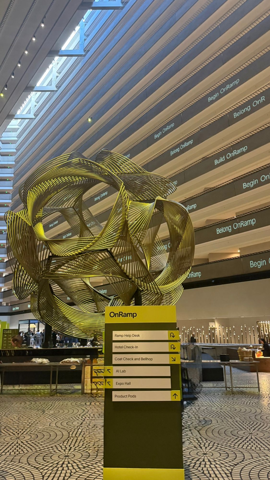 The OnRamp 2026 atrium in San Francisco: a gold ribbon sculpture beside the event's wayfinding sign.